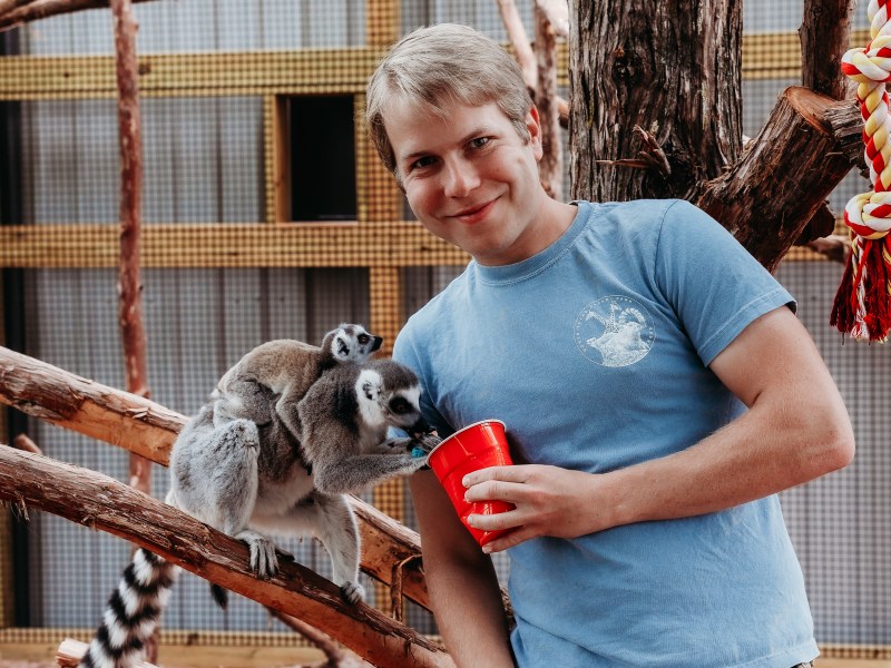 Man in blue shirt stands by lemurs on tree branches, holding a red cup.