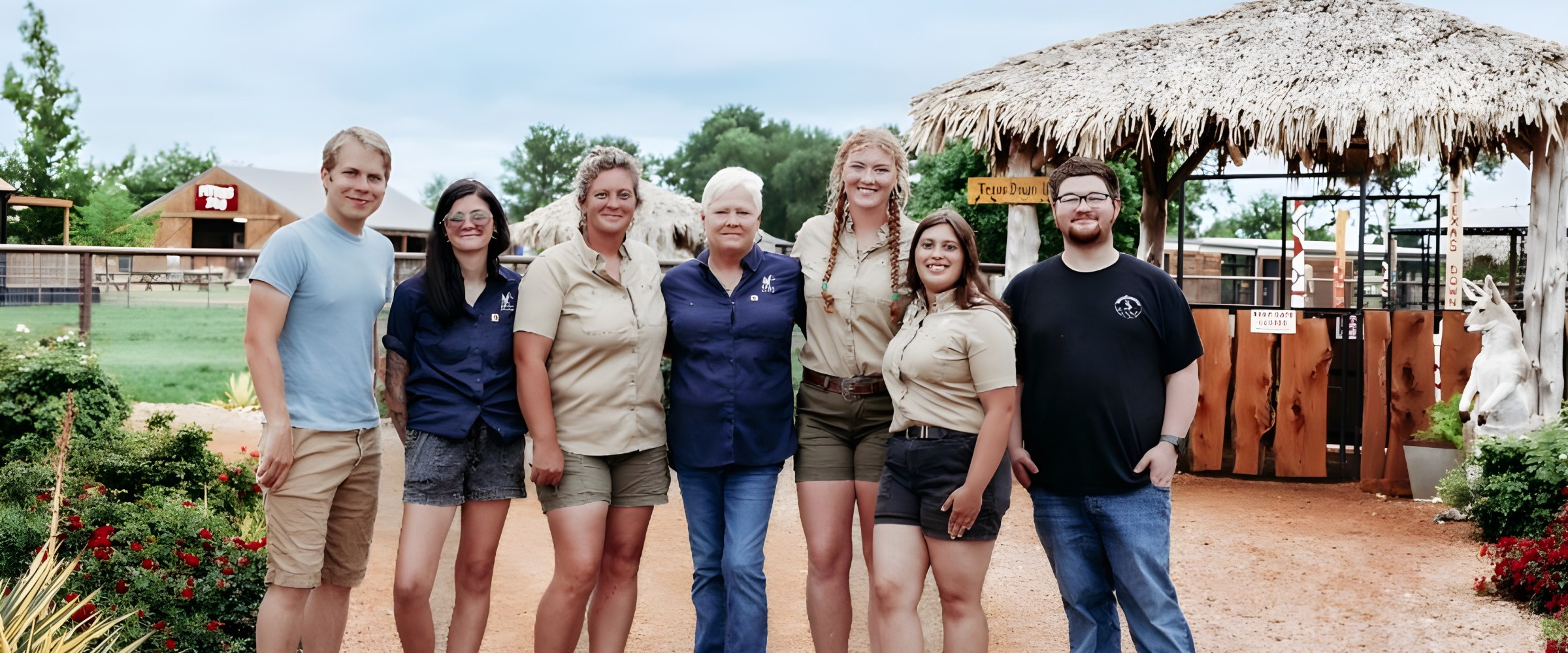 Seven people stand smiling outdoors near a thatched-roof structure.